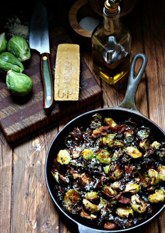 air fried Brussels Sprouts with Bacon in cast iron skillet. Cutting board with knife, brussels, parm wedge and bottle of olive oil behind.