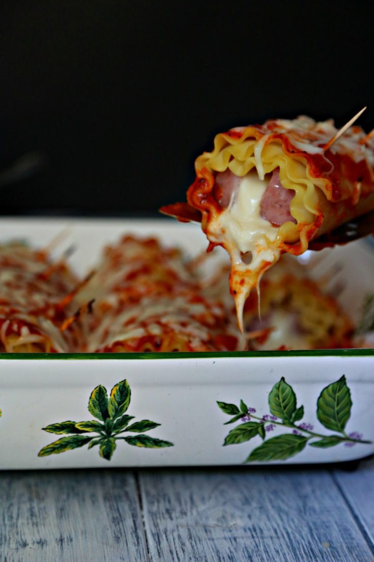 Kielbasa Lasagna rolls in casserole dish. Serving spoon pulling one roll out.