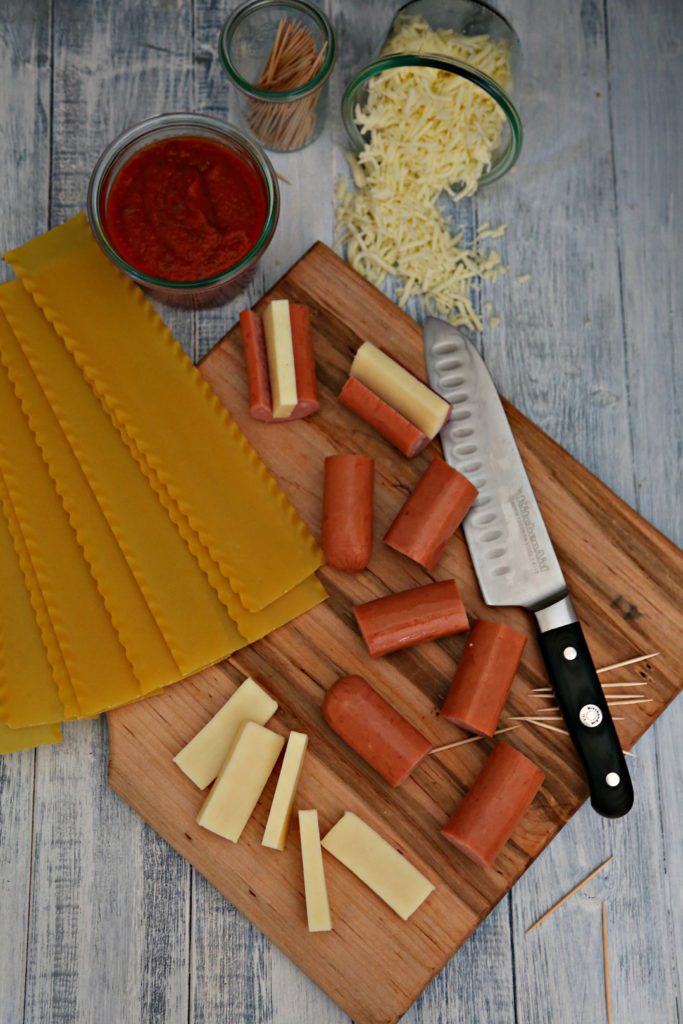 sliced pieces of kielbasa sausage on cutting board with knife, toothpicks and cheese slices. Lasagna noodles, small glass jar of sauce, small glass jar of toothpicks, small glass jar with shredded cheese spilling out in background. 