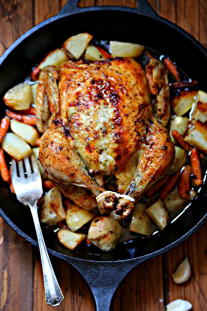 closeup of roasted chicken in cast iron skillet with potatoes and carrots. Fork to left of chicken. 