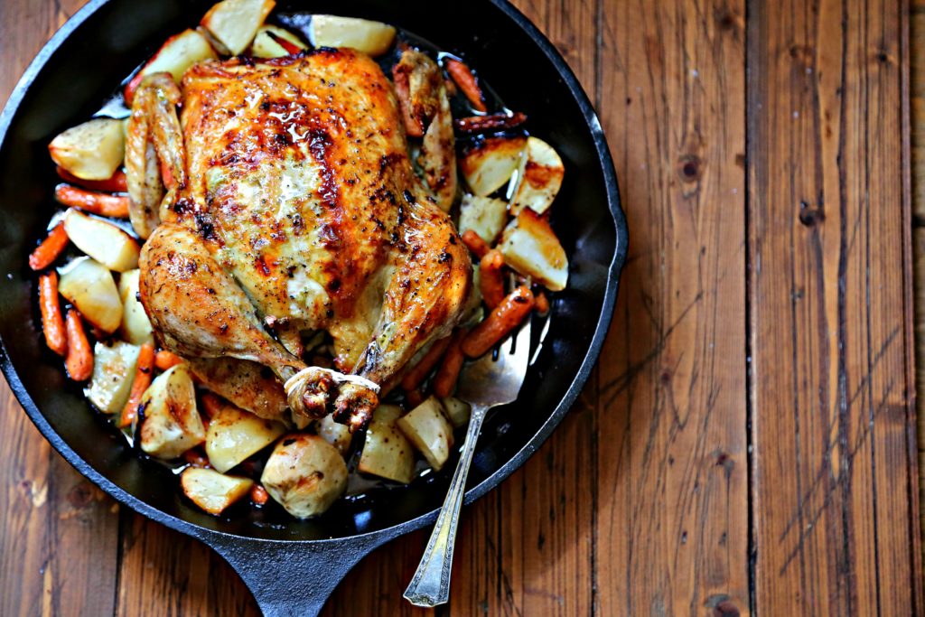 Roasted chicken in cast iron skillet with potatoes, carrots and serving fork. 