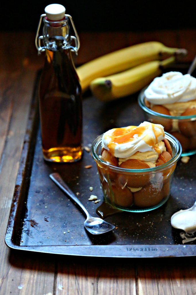 2 glass jars of banana pudding. Topped with whipped cream and caramel sauce sitting on baking sheet. Spoons, bottle of vanilla extract and bananas to side.