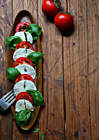Slices of tomatoes, mozzarella and fresh basil on brown oval tray. Serving fork to side and tomatoes in background.