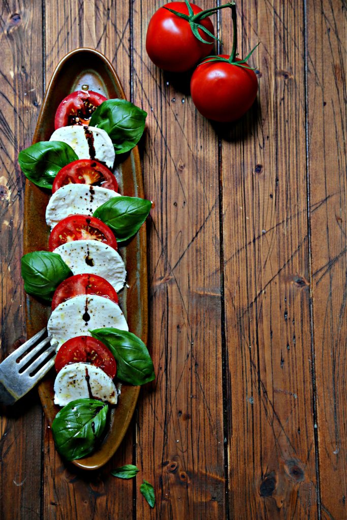 Slices of tomatoes, mozzarella and fresh basil on brown oval tray. Serving fork to side and tomatoes in background.