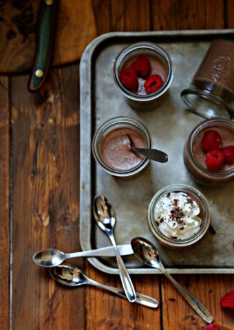 Chocolate Amaretto Panna Cotta in mini glass jars. Some topped with raspberries, others with whipped cream and chocolate shavings. Small spoons to side.