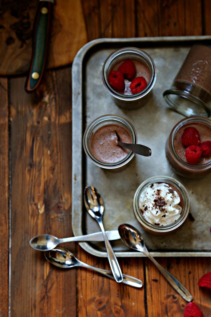 Chocolate Amaretto Panna Cotta in mini glass jars. Some topped with raspberries, others with whipped cream and chocolate shavings. Small spoons to side.