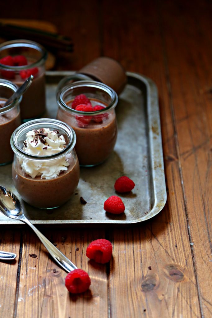 Chocolate Amaretto Panna Cotta in small glass jars. Garnished with whipped cream chocolate shavings, raspberries. Small spoons to side.