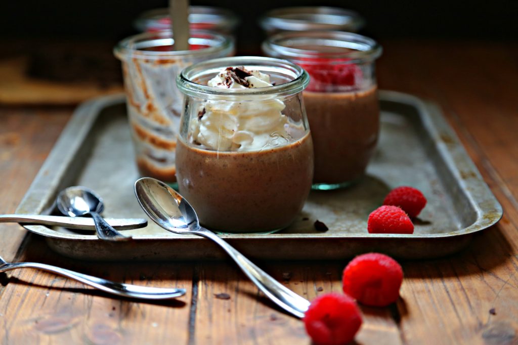 Chocolate Amaretto Panna Cotta in glass jars. Garnished with whipped cream, chocolate shavings and raspberries. Small spoons to side.