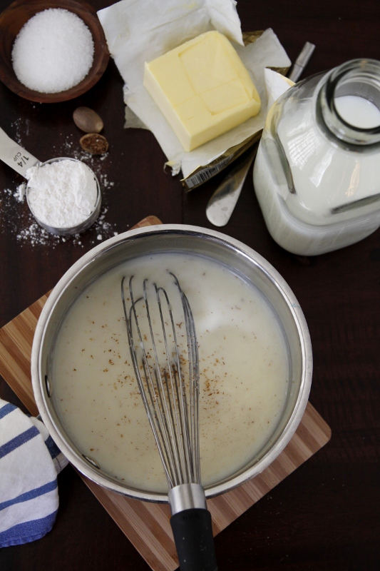 sauce pan with bechamel sauce with whisk. Glass jar of milk, butter and measuring spoons to side.