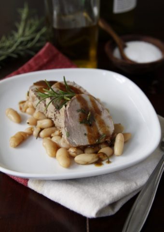 white plate with cannelli beans topped with slices of pork tenderloin and sauce.
