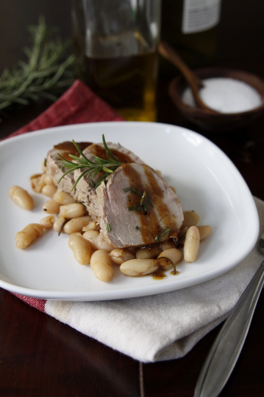 White plate with cannellini beans topped with pork tenderloin slices and sauce. Napkin under plate. Salt cellar, olive oil jar and rosemary blurred in background. 