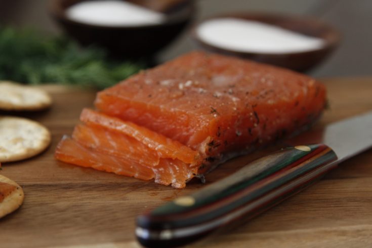 gravlax on cutting board with knife. Small brown bowls of salt and sugar in background.
