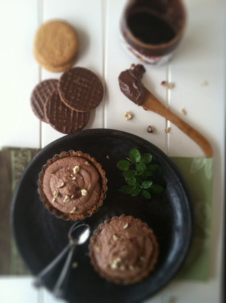 2 nutella mascarpone chocolate tarts on brown plate with small spoons. Cookies, and jar of nutella with wooden spoon behind.