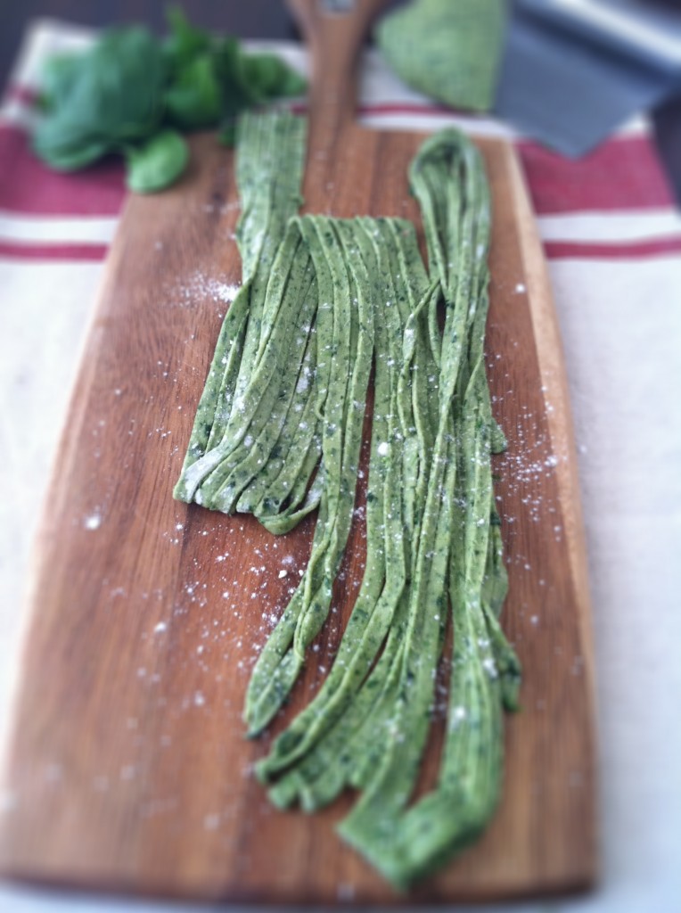 homemade spinach pasta on a wooden board beside fresh spinach and spinach dough on a pastry cutter