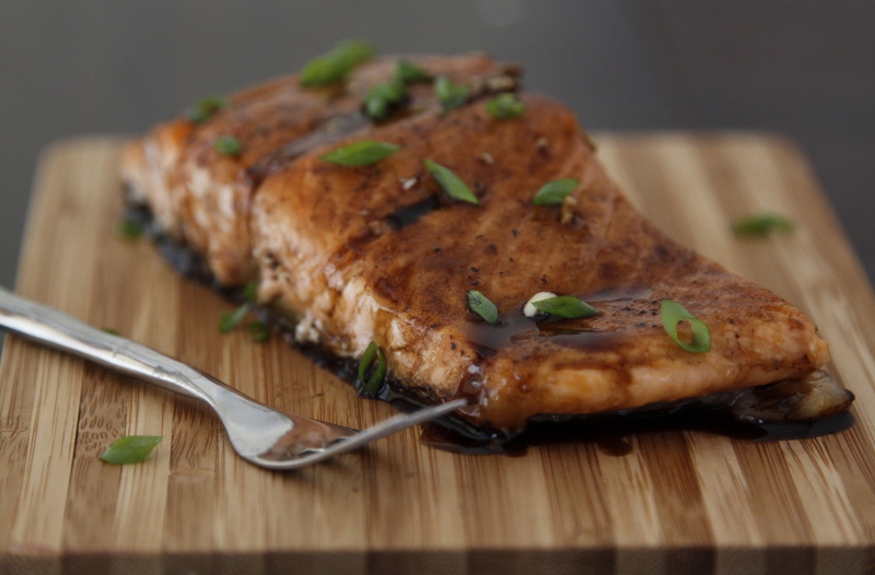 Piece of Maple Balsamic Glazed Salmon on cutting board garnished with green onions. Fork in front. 