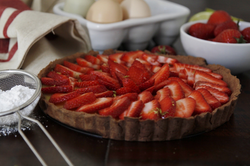 strawberry tart surrounded by napkin, sieve with powdered sugar, container or eggs and bowl of strawberries. 