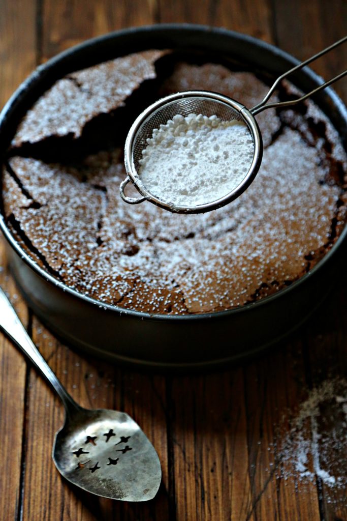 chocolate cake with metal sifter of powdered sugar above. Serving piece to side. 