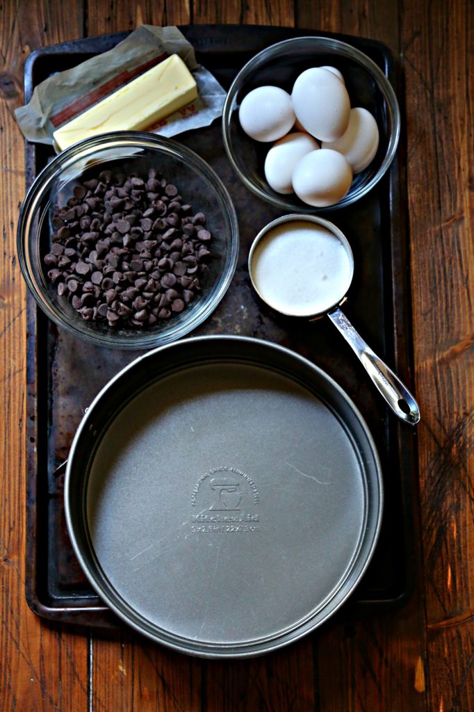 baking sheet with ingredients for chocolate cake. Glass bowl of chips, bowl of eggs, measuring cup with sugar, butter, and pan.
