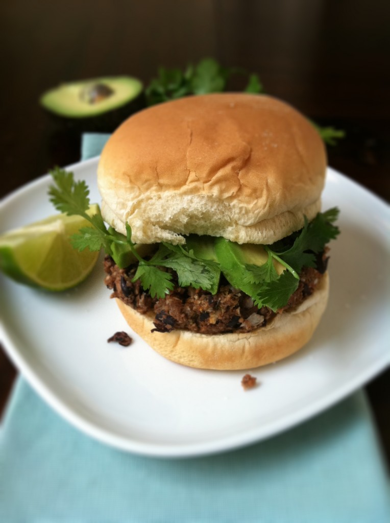 Black Bean Veggie Burger on white plate. Lime wedge to side. Blue napkin under plate. Avocado half in background. 