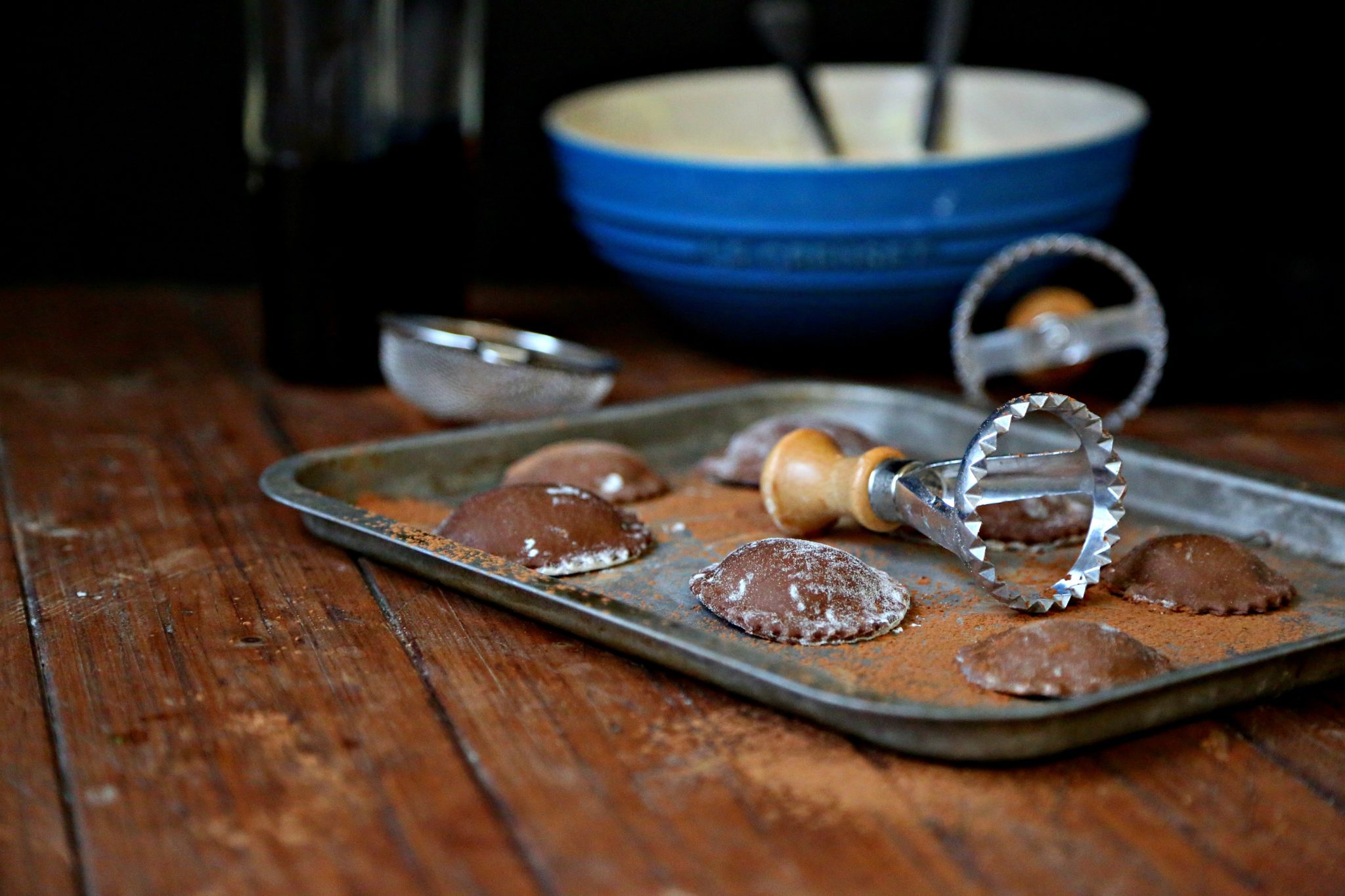 chocolate ravioli on baking sheet with ravioli stamps and blue bowl of filling in background.