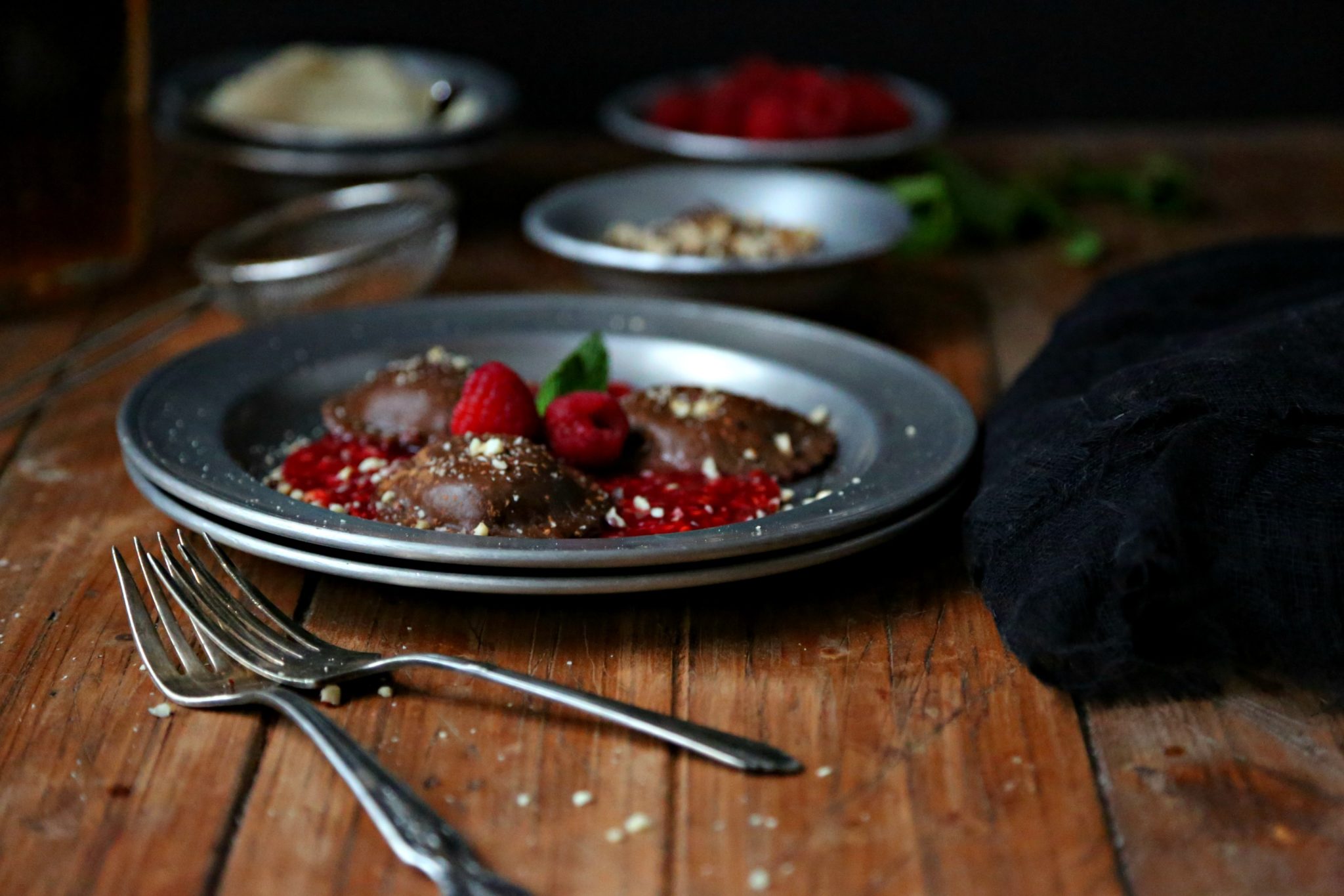 chocolate ravioli on a silver plate on top of raspberry puree with 2 forks, topped with mint, and fresh raspberries