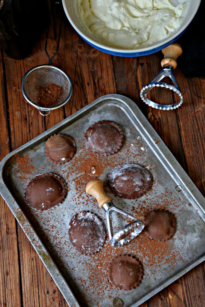 chocolate ravioli on a baking tray surrounded by ravioli stamps and bowl of filling.
