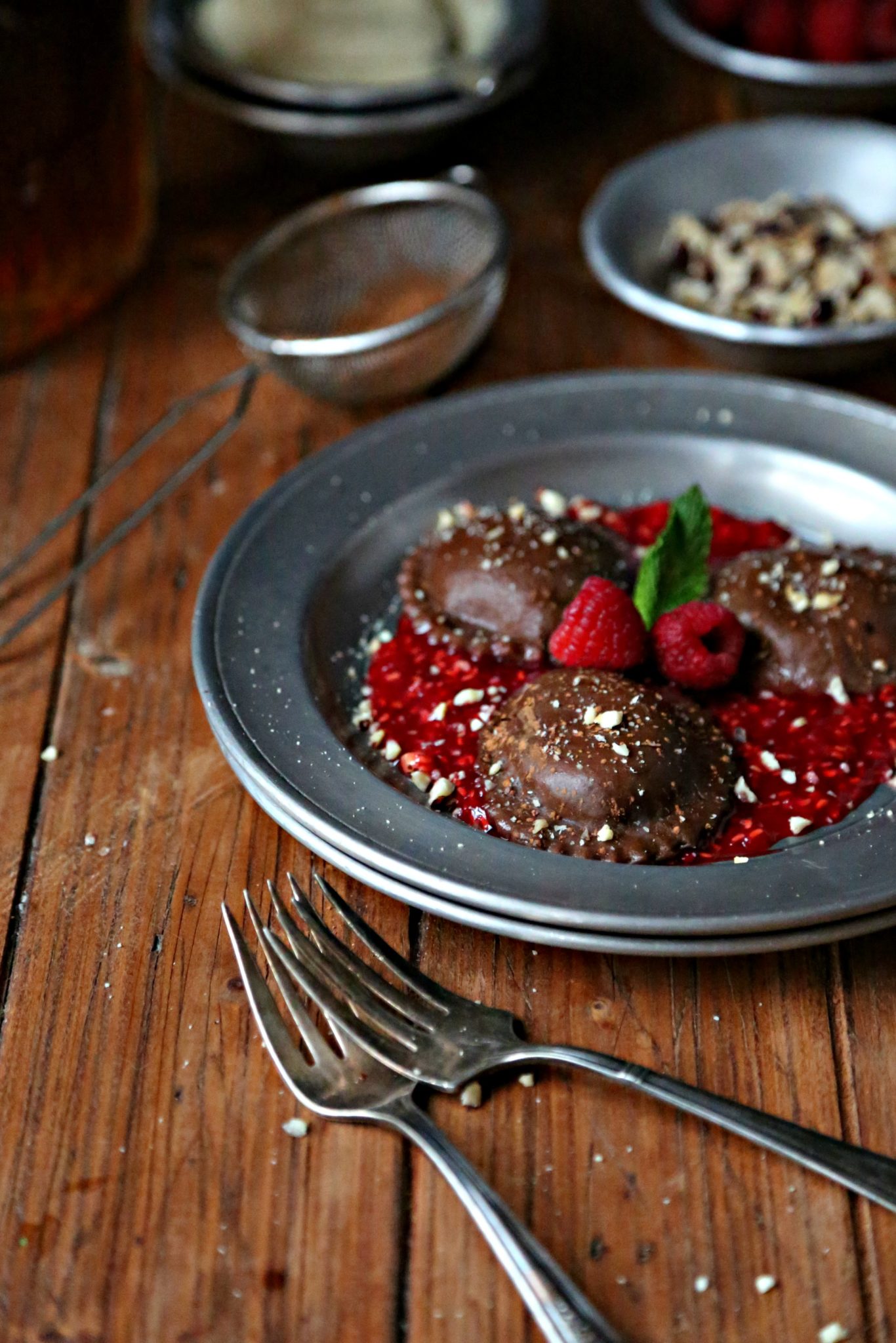 chocolate ravioli on a silver plate with raspberry puree and chopped nuts with 2 silver forks. Small silver bowls of nuts, raspberries and a small sifter with cocoa powder in background.