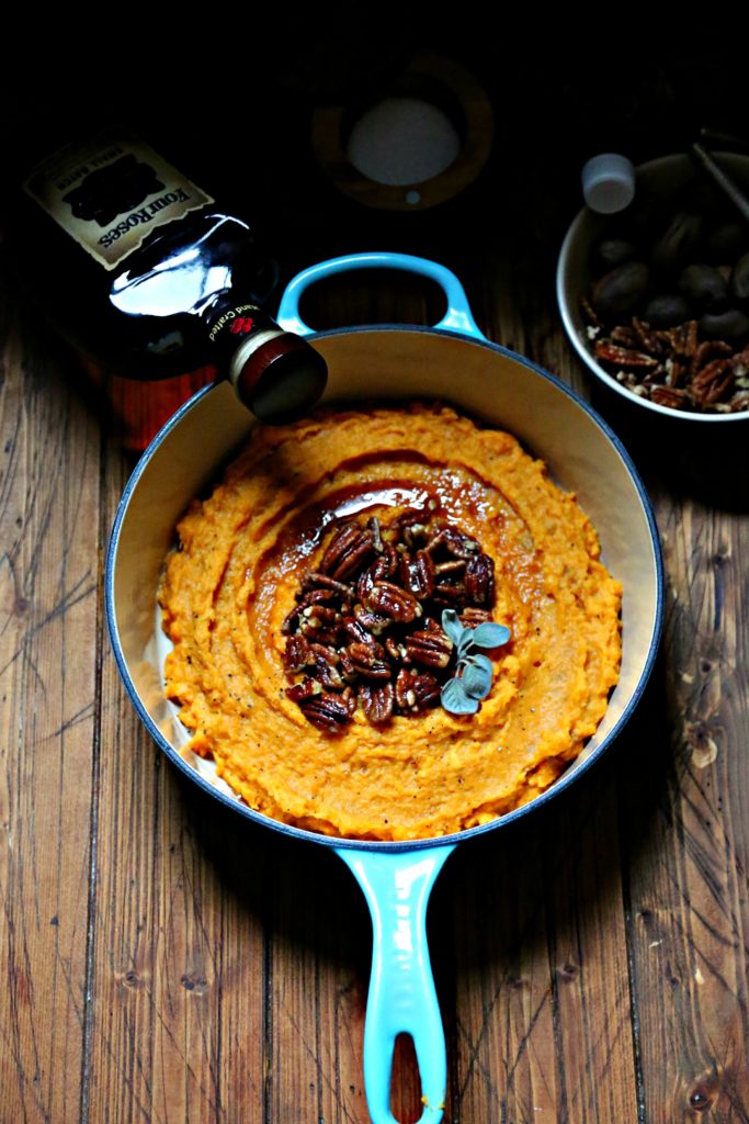 Boozy Smashed Sweet Potatoes in blue pot with bowl of pecans and bottle of bourbon to side