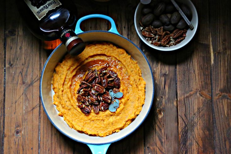 Boozy Smashed Sweet Potatoes in blue pot with bowl of pecans to side and bottle of bourbon