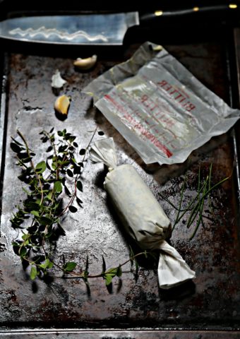 compound butter in parchment, fresh herbs, butter wrapper and garlic cloves on baking sheet.