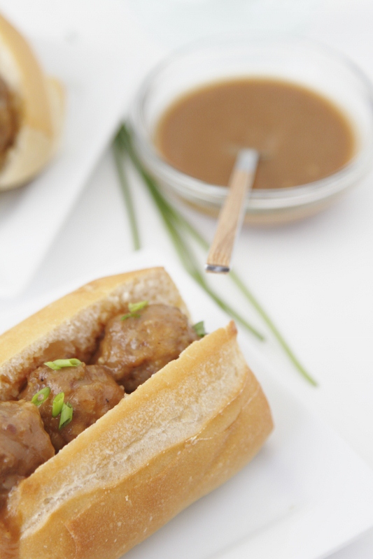 mini meatball sub garnished with chives on white plate. Small glass bowl of sauce with spoon in background. 
