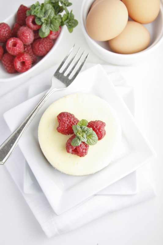 Mini cheesecake on white plate with fork. Garnished with mint and raspberries. White bowl of raspberries, white bowl of eggs in background. 