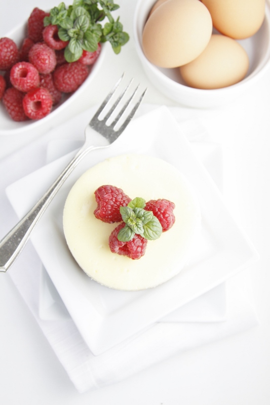 mini cheesecake on white plate garnished with mint and raspberries. White bowl of raspberries and eggs in background.