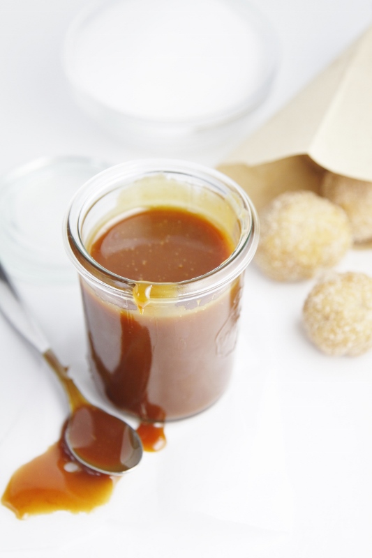 small glass jar of caramel sauce with spoon with sauce in front of jar. Bag of doughnut holes in background.