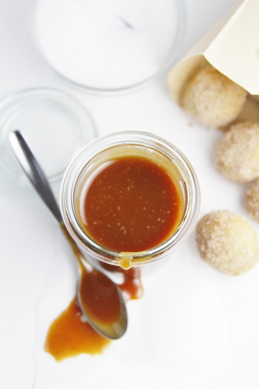 small glass jar of caramel sauce. Spoon with sauce in front of jar. Bag of doughnut holes in background. 