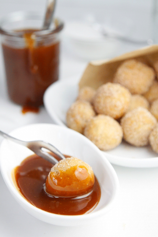 Small white bowl of caramel sauce. Doughnut hole on fork being dipped into sauce. Bag of doughnuts and small glass jar of caramel sauce in background. 