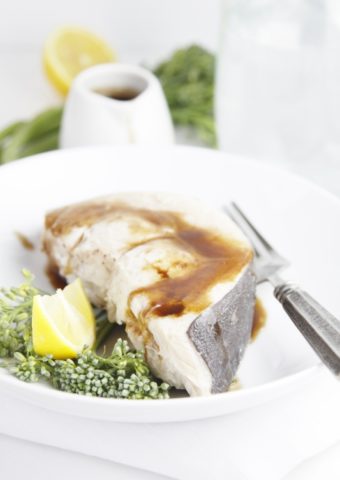 swordfish with sauce, broccoli and lemon wedge on white plate with fork. Small white pitcher of sauce in background.