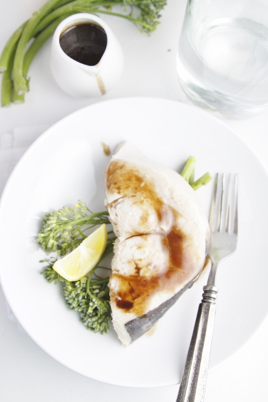 Swordfish with Balsamic Brown Butter Sauce on white plate with broccolini, lemon wedge and fork. Small pitcher of sauce and broccolini in background. 