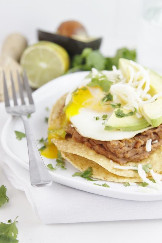 Stacked Breakfast Tostada topped with runy egg, avocado slices and cheese on white plate with fork. Blurred ingredients in background. 