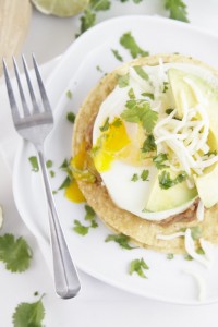 Stacked Breakfast Tostada topped with runy egg and grated cheese on white plate with fork. Cilantro around plate. 