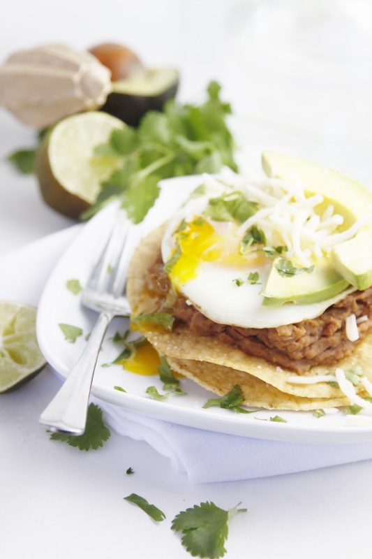 Stacked Breakfast Tostada topped with runy egg and avocado slices on white plate with fork. Blurred ingredients in background. 
