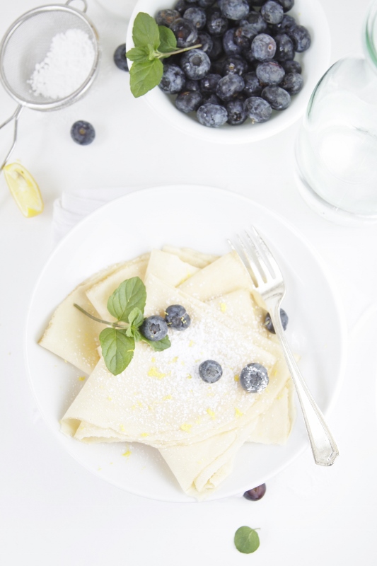 Lemon Crepes with Blueberries on white plate with fork. White bowl of blueberries, metal sifter with powdered sugar and glass carafe in background. 