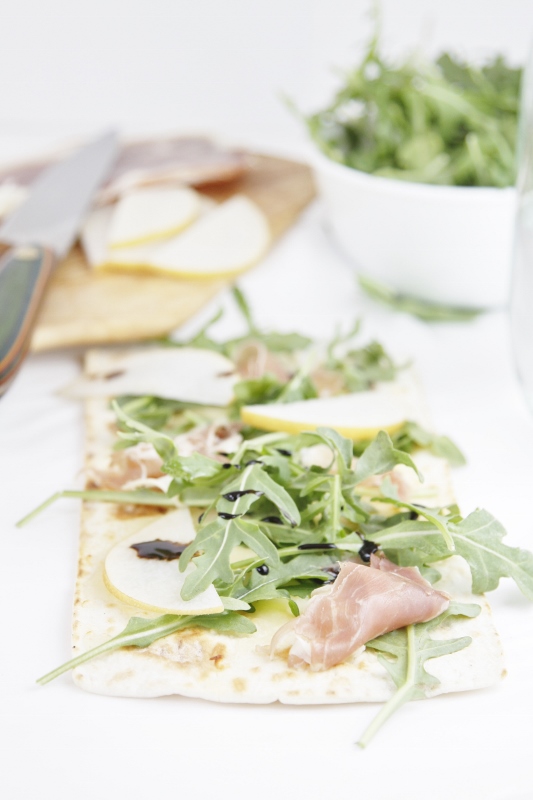 Prosciutto Pear and Arugula Flatbread Pizza. Cutting board with knife, pear slices in background. White bowl with arugula to right. 