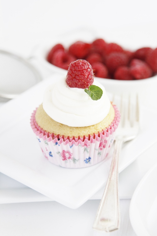 Cupcake garnished with raspberry and mint on white plate with fork. White bowl of raspberries in background. 