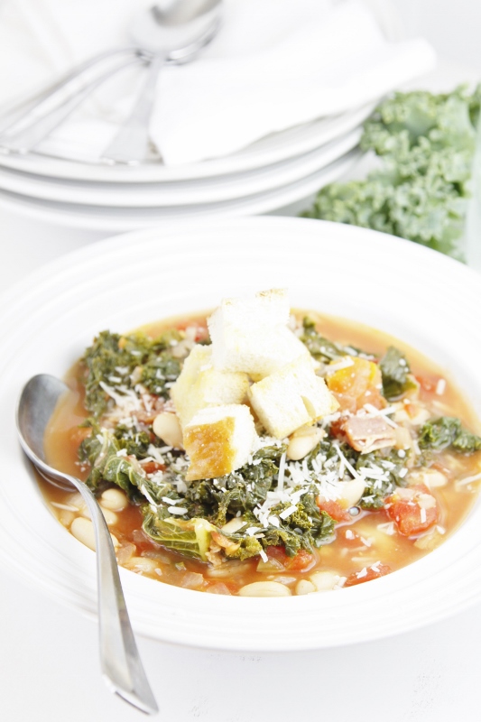 Tuscan Soup in white bowl with spoon. Stack of white bowls with spoons in background with leaf of kale to side. 