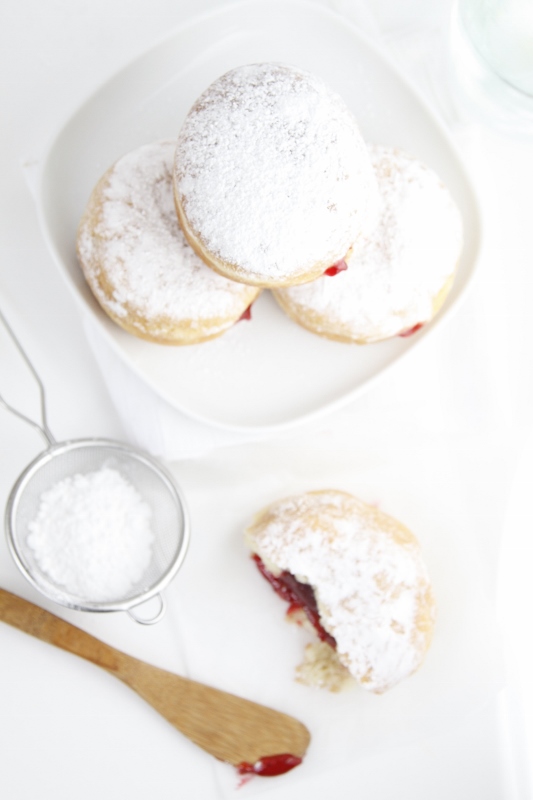 Strawberry Filled Doughnuts stacked on white plate. Metal sifter with powdered sugar, half doughnut with filling showing and wooden knife in front. 