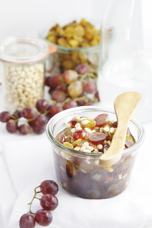 Raisin and Nut Agrodolce in small glass jar with wooden spoon laying across top. Glass jar of pine nuts, glass jar of golden raisins, red grapes and glass carafe in background.