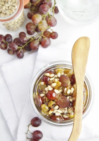 Glass jar of agrodolce with wooden spoon laying across. Red grapes and glass jar of pine nuts in background.