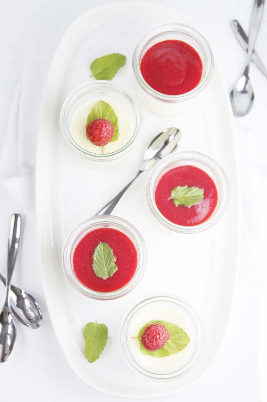 5 small glass jars of raspberry panna cotta on white tray. Small spoons and fresh mint scattered around.