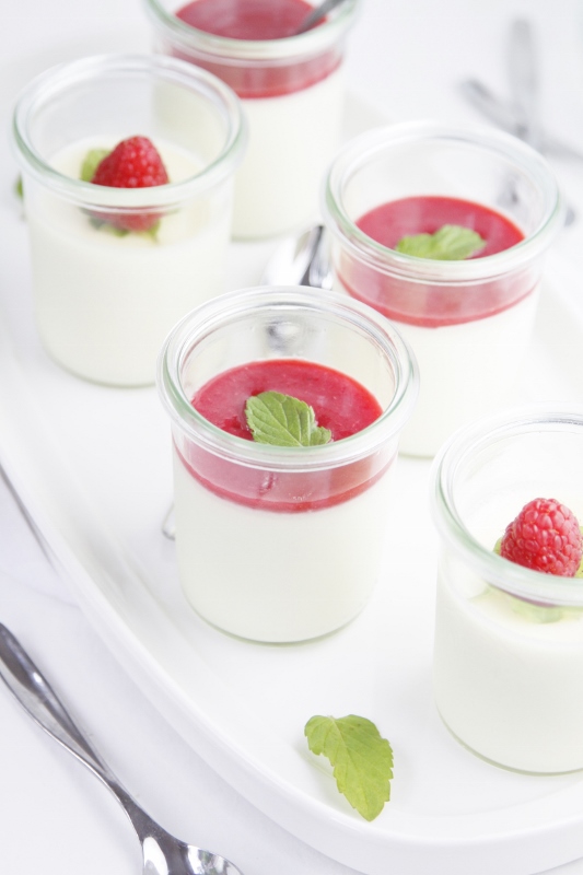 5 small glass jars of Raspberry Panna Cotta on white tray. Small serving spoons scattered around.